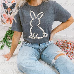 May include: Dark gray t-shirt featuring a rabbit silhouette made of white pearls. The shirt is paired with light blue ripped jeans. The image is taken indoors.