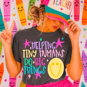 May include: A charcoal gray t-shirt featuring the colorful text "HELPING TINY HUMANS DO BIG THINGS" in purple, yellow, pink, and teal, accompanied by pink star graphics and a large yellow smiley face. The shirt is worn with a multi-colored trucker hat.