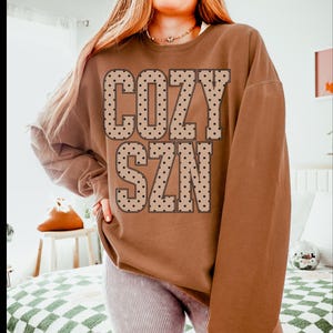 Peut inclure: Un sweat-shirt marron à manches longues et col rond avec l'inscription COZY SZN en grandes lettres capitales. Les lettres sont beige clair avec un motif à pois noirs et un fin contour noir. Le sweat-shirt a une coupe décontractée.