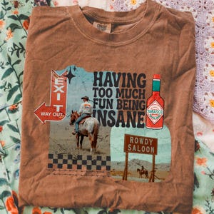 May include: Brown t-shirt with a graphic featuring a cowboy on a horse, a Tabasco bottle, and the text "HAVING TOO MUCH FUN BEING INSANE." The design includes an "EXIT" sign and a "ROWDY SALOON" sign.