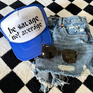 May include: A blue and white trucker hat with the text "be savage not average" in black. The hat is on a black and white checkered surface with a pair of black sunglasses and blue denim shorts.