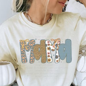 May include: Cream-colored t-shirt with the word "Mama" in a decorative font. The letters are adorned with fall-themed patterns, including pumpkins, flowers, and polka dots. A gold chain necklace is visible.