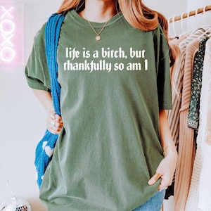 Puede incluir: Camiseta verde oliva con el texto blanco "life is a bitch, but thankfully so am I" en fuente gótica. La camiseta es de corte holgado y tiene mangas cortas. Se ve un bolso azul tejido.