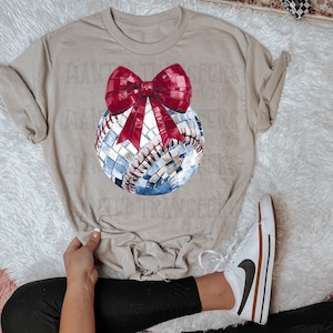 May include: A beige t-shirt with a baseball-shaped disco ball with a red bow on top. The shirt is being worn by a person with black pants and white sneakers.