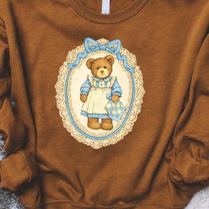 May include: Brown sweatshirt featuring a vintage-style illustration of a teddy bear. The bear is dressed in a blue and cream-colored dress and holds a small blanket. The illustration is framed by a blue ribbon and a decorative lace-like oval.
