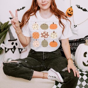 Può includere: T-shirt bianco con una griglia di nove zucche illustrate in diversi colori e motivi. Le zucche presentano disegni floreali, a pois e a tinta unita. La maglietta è indossata da una persona.