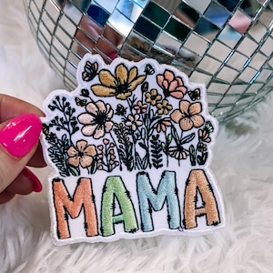 May include: Embroidered patch with the word "MAMA" in orange, green, and blue, beneath a floral design with yellow and peach flowers. The patch is on a white background with a fluffy texture.