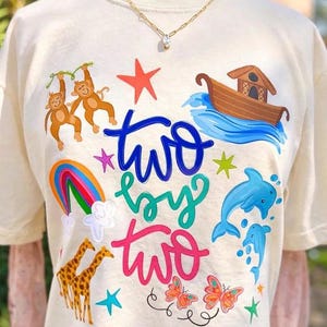 May include: Cream-colored t-shirt with a colorful graphic design. The design includes the words "two by two" in blue, green, and pink, along with illustrations of monkeys, a rainbow, giraffes, dolphins, butterflies, a boat, and stars.