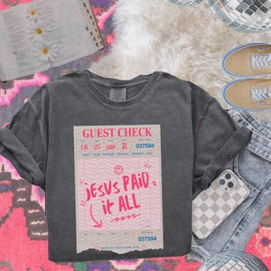 May include: A dark gray t-shirt with a vintage-style guest check design. The check says "Jesus Paid it All" with a smiley face and a check mark. The check number is 037594.