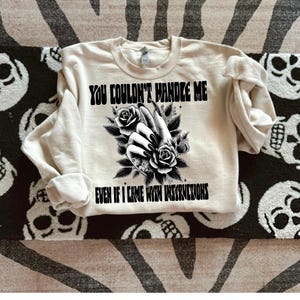 May include: A cream colored sweatshirt with black text that reads "You couldn't handle me even if I came with instructions". The text is surrounded by a black and white illustration of a hand with long nails and flowers.