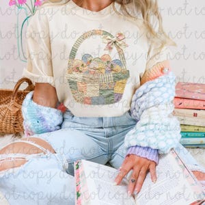 Puede incluir: Camiseta color crema con un diseño de cesta de Pascua llena de huevos decorados. La cesta tiene un asa verde y un lazo rosa. La camiseta forma parte de una colección con temática de primavera o Pascua.