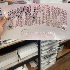May include: A light pink heat transfer vinyl sheet with the word "TEACH" in a rhinestone design, and the words "TEACH-LIVE-INSPIRE" below. The design includes rhinestone flower accents. The sheet is held up by a hand.