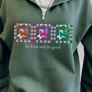Könnte beinhalten: Olivgrünes Sweatshirt mit Viertelreißverschluss und farbenfrohem Schmetterlingsdesign. Das Design zeigt drei quadratische Tafeln mit Schmetterlingen, Herzen und Sternen. Der Text "be kind and do good" ist darunter gedruckt.