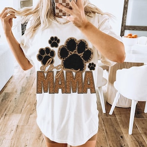 May include: White t-shirt with a brown and black paw print design and the text "dog mama" in gold glitter.