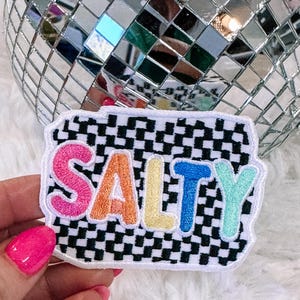 May include: Embroidered patch with the word "SALTY" in colorful letters. The letters are outlined in white and sit on a black and white checkered background. The patch has a white border.