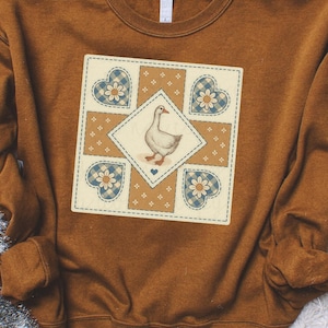 May include: A brown sweatshirt featuring a central design of a goose within a diamond shape. The design is surrounded by four heart shapes with blue gingham and floral accents. The sweatshirt has a crew neck and long sleeves.