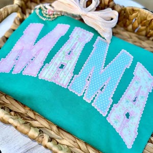Puede incluir: Camiseta de color turquesa con la palabra "MAMA" en letras grandes y decorativas. Las letras tienen patrones rosas y azules, incluyendo diseños florales y de lunares. Un lazo rosa claro y adornos decorativos en la parte superior.