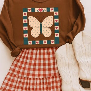 May include: A brown sweatshirt featuring a butterfly design, combined with a red and white gingham skirt and white cowboy boots. The butterfly is cream with blue cross details, framed by a green border with red hearts. The boots have brown heels.
