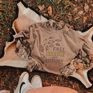 May include: A tan short-sleeve t-shirt layered over a long-sleeve camouflage shirt. The t-shirt features a graphic of swirling autumn leaves and the text "FALL BREEZE AND AUTUMN LEAVES" in yellow, orange, and dark brown lettering on the front.
