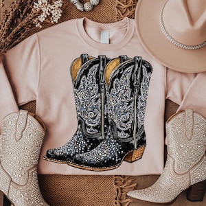 May include: A light pink sweatshirt featuring a pair of black cowboy boots with intricate rhinestone detailing. The boots have a pointed toe and a stacked heel. A beige cowboy hat is visible in the background, along with a pair of rhinestone-covered cowboy boots.