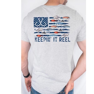 Pode incluir: Uma camiseta cinza com um design de bandeira americana desbotada com iscas e anzóis de pesca. O texto "Keepin' It Reel" é impresso abaixo da bandeira.