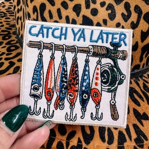 May include: A white patch with the text "CATCH YA LATER" in blue, featuring fishing lures and a fishing rod. The patch is set against a leopard print background.