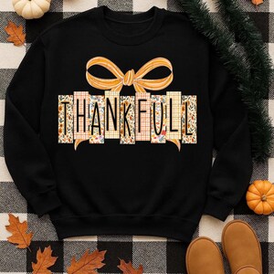 May include: Black sweatshirt with the word "THANKFUL" in a decorative design. The word is made up of vertical rectangles with floral and plaid patterns. A large orange bow is above the word. The sweatshirt is surrounded by fall-themed decorations.