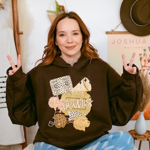 May include: A brown hoodie featuring a graphic design with the word "Bears" in a script font. The design includes a football helmet, a bear head, a megaphone, and a scoreboard. The hoodie has a drawstring hood.