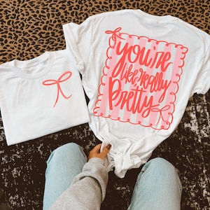May include: Two white t-shirts. One has a red bow graphic, the other has the text "You're like really pretty" in red script on a pink striped background. The shirts are on a leopard print surface.