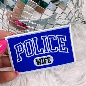 May include: A blue and white embroidered patch with the words "POLICE WIFE". The patch has a white border and is rectangular in shape. A disco ball is in the background.