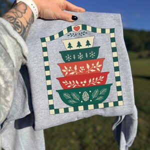 May include: A gray sweatshirt featuring a stacked bowl design in red, green, and white. The bowls are decorated with Christmas tree, snowflake, and botanical patterns. The design is framed with a green and white checkered border and a heart.