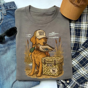 Pode incluir: Camiseta cinza com um golden retriever usando um boné "GOOD BOY" e segurando um pato. O cão está sentado ao lado de um cooler camuflado com o texto "YETI" e "DUCK LIFE". Um boné marrom, óculos de sol e jeans também estão na imagem.