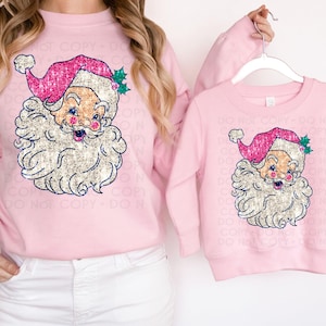 May include: Pink Christmas sweatshirts with a sparkly glitter Santa Claus design. The larger sweatshirt has long sleeves and the smaller sweatshirt has short sleeves.