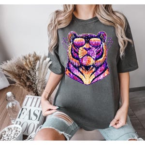 May include: A dark gray t-shirt with a glittery purple and orange tiger wearing sunglasses. The tiger is in a geometric design.