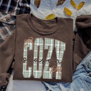 Puede incluir: Una sudadera de cuello redondo color marrón oscuro con la palabra COZY en letras grandes de bloque a cuadros verde claro y blanco con un borde de estampado de leopardo. La palabra season está escrita en cursiva marrón en el centro, con dos lazos naranjas.