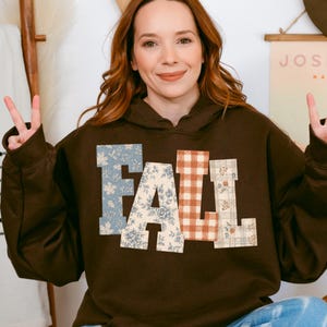 May include: A dark brown hooded sweatshirt featuring the word FALL in large block letters across the chest. Each letter has a different pattern: the F is blue floral, the A is cream floral, the L is orange gingham, and the L is plaid.