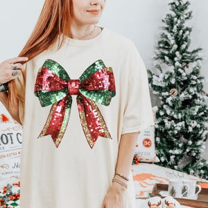 以下が含まれることがあります： クリーム色のTシャツ。大きなスパンコールのリボンデザインが特徴です。リボンは赤、緑、金のきらびやかなスパンコールの組み合わせで、きらめく効果を生み出しています。ゆったりとしたフィット感で、普段着に適しています。背景にはクリスマスツリーがあります。