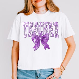 May include: White t-shirt with the word "TEACHER" in purple floral print and a large purple bow. The shirt is a casual style, suitable for everyday wear. The design is centered on the front of the shirt.