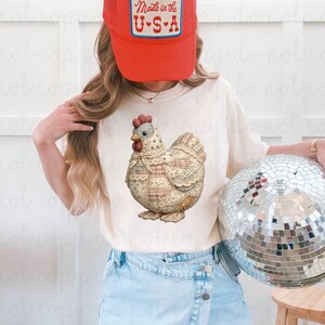 May include: A cream-colored t-shirt featuring a detailed illustration of a patchwork chicken. The model is wearing a red baseball cap with a "Made in the U.S.A." patch. The model is also holding a disco ball.