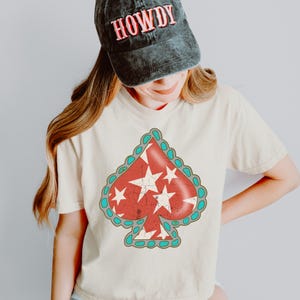May include: Cream-colored t-shirt featuring a distressed red spade design with white stars and a turquoise border. A black cap with the word "HOWDY" in red is worn on the head.