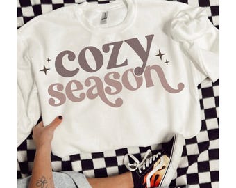 May include: White crewneck sweatshirt with the words "cozy season" in brown and mauve lettering. The sweatshirt is on a black and white checkered surface. A person is wearing denim shorts and black sneakers with flames.