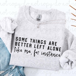 Puede incluir: Sudadera gris claro con el texto negro "SOME THINGS ARE BETTER LEFT ALONE like me, for instance". Se ve un collar plateado con un pequeño colgante. La sudadera está doblada sobre unos vaqueros azules.