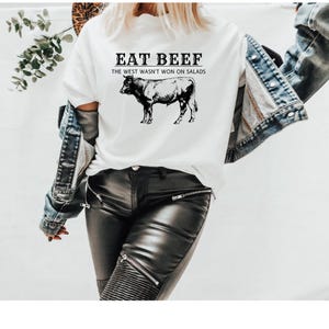 Puede incluir: Camiseta blanca con texto negro que dice "EAT BEEF - THE WEST WASN'T WON ON SALADS". Debajo del texto hay una ilustración en blanco y negro de una vaca. La persona que lleva la camiseta también lleva una chaqueta vaquera y pantalones de cuero negro.