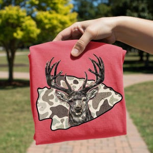 May include: A folded red t-shirt featuring a detailed illustration of a deer with large antlers. The deer is set against a camouflage-patterned background. The shirt is held by a hand.