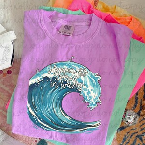 May include: A lilac Comfort Colors t-shirt with the phrase "Happiness comes in waves" and a blue and white wave graphic. Other colorful t-shirts are visible in the background. The shirt has a crew neck and short sleeves.