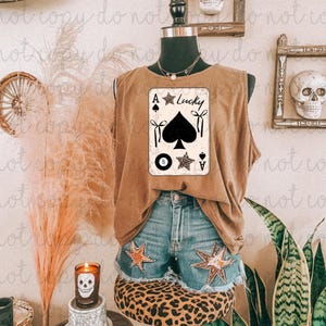 Op de afbeelding: Een bruine tanktop met een speelkaartafbeelding met de tekst "Lucky" en een schoppen, een achtbal en sterren. De tanktop wordt gecombineerd met een spijkerbroek met sterrenpatches. De mannequin staat op een kruk met luipaardprint.