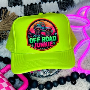 Könnte beinhalten: Neongrüne Trucker-Cap mit Mesh-Rückseite und gebogenem Schirm. Auf der Vorderseite befindet sich ein kreisförmiger Aufnäher mit einer Geländewagen-Grafik und dem Text "OFF ROAD JUNKIE" in Rot, Grün und Orange.