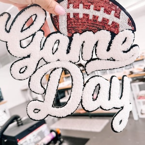 May include: A decorative patch featuring the text Game Day in white fuzzy chenille lettering with a black outline. Above the text is a football shape covered in shimmering red and white sequins. The entire piece has a black border.