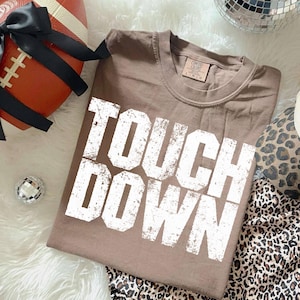 May include: A brown t-shirt with the words "TOUCH DOWN" in large, distressed white letters. The shirt is folded and lies on a white, fluffy surface, near a football and leopard print fabric. A small disco ball is also present.