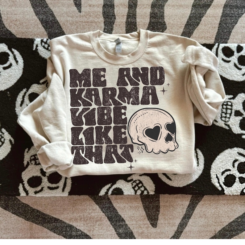 May include: A beige sweatshirt with the text "Me and Karma Vibe Like That" in black, with a skull graphic with heart-shaped eyes.